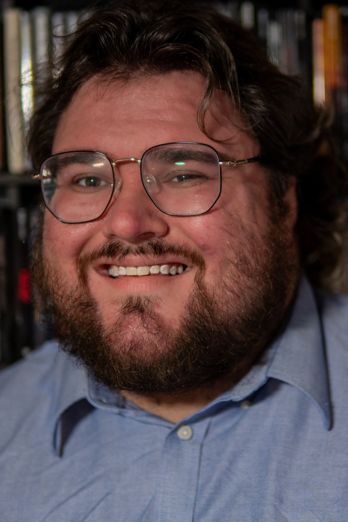 Portrait of Tanner Richard Craft, wearing glasses and a blue button-down shirt, in front of shelves of films